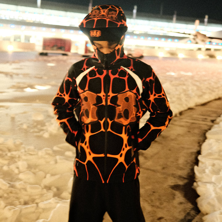 Person wearing a black and orange patterned outfit standing against a dark background with lights.