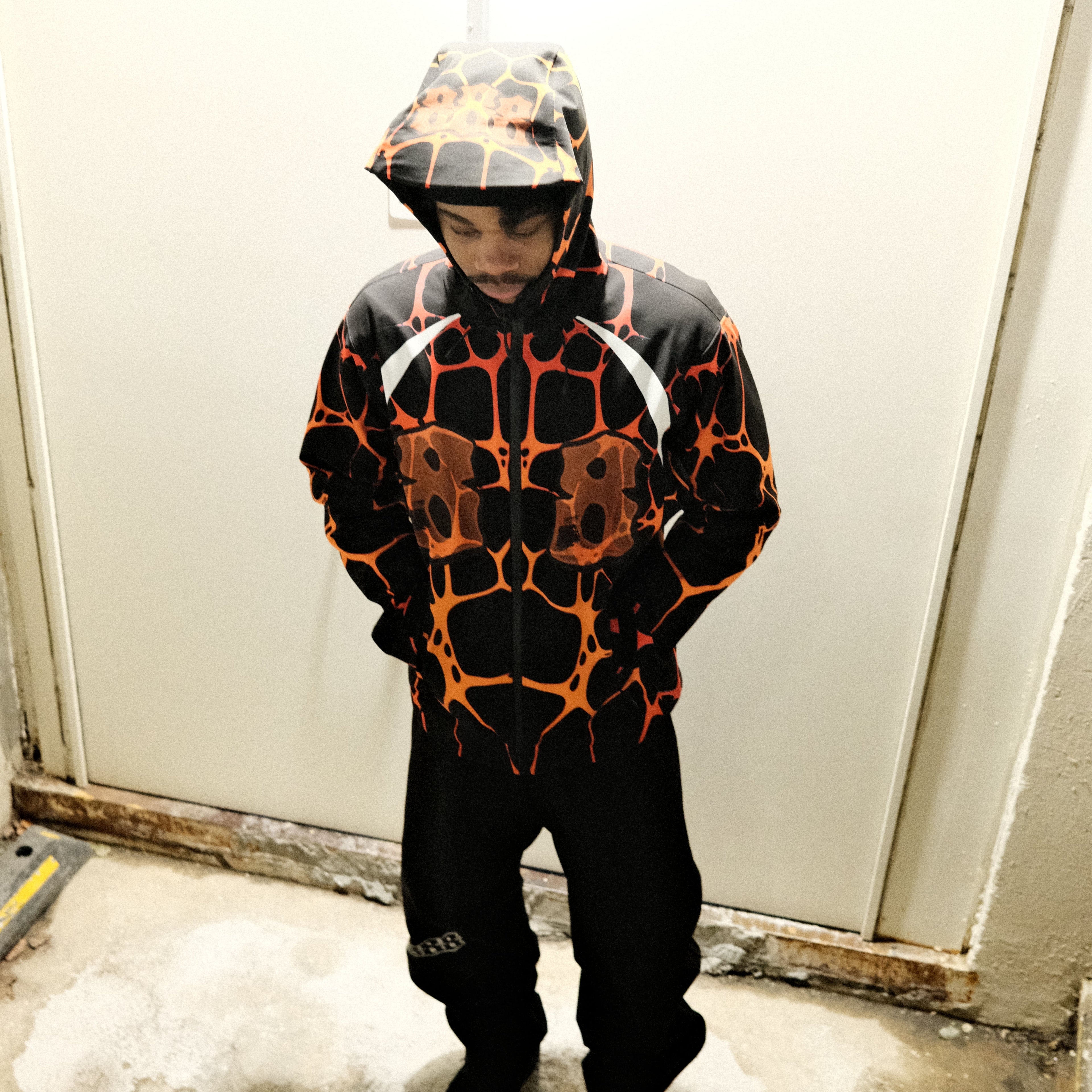 Person wearing a black and orange patterned jacket standing in front of a white door.